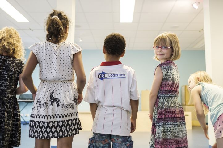 Fünf  Kinder stehen in einer Reihe mit dem Rücken zur Kamera, ein Mädchen dreht sich um und lächelt in die Kamera